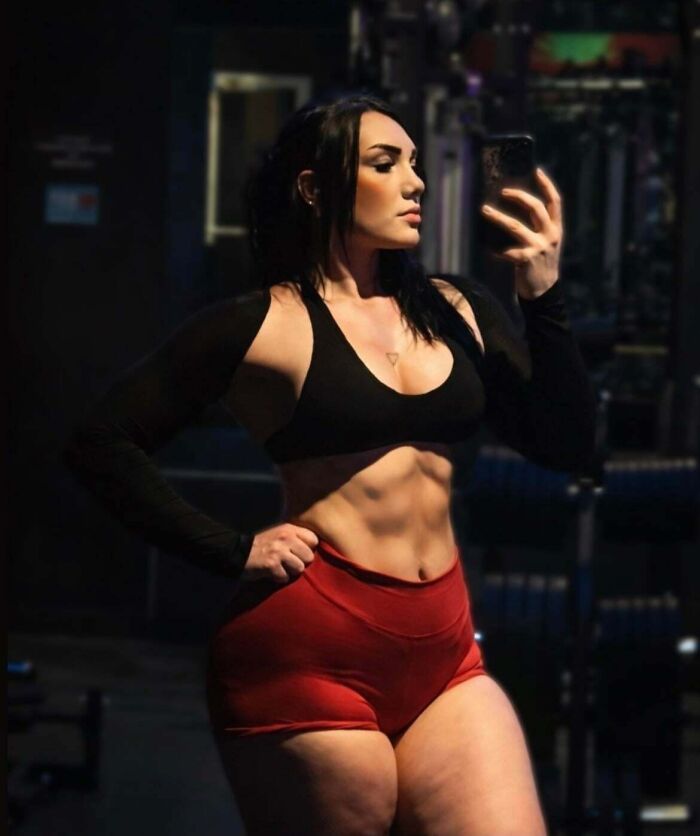 A woman in a sports bra and red shorts poses for a natural social media photo in a gym.