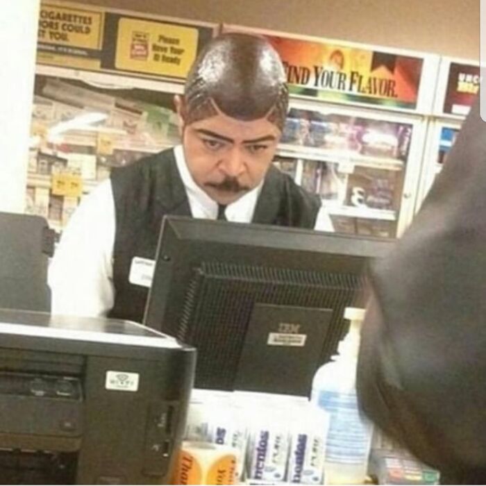 A man with unique, bold hairstyles and drawn-on eyebrows working behind a counter.
