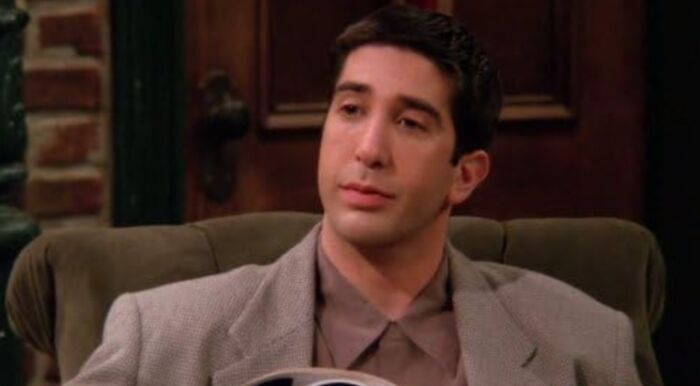 Close up of Ross Geller from Friends in a brown shirt and gray jacket, answering about his academic field.