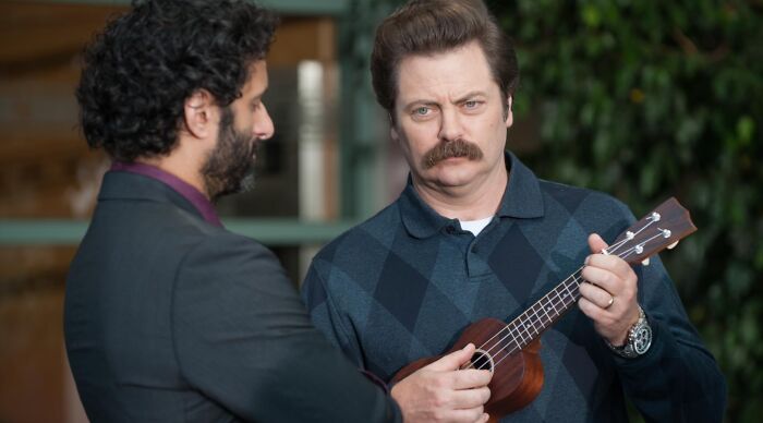 Ron Swanson's job title in Parks and Recreation: Ron plays a ukulele while looking annoyed as a man looks at him.