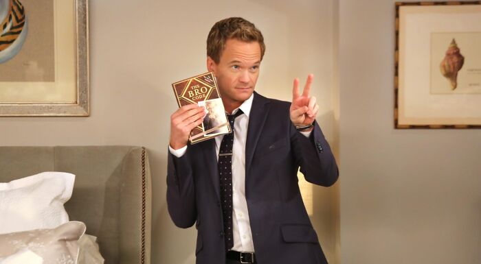 Barney Stinson in a suit, holding The Bro Code book, making a peace sign for a TV show character quiz.