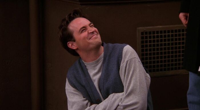 Chandler Bing from Friends, looking upward with a pleased expression, wearing a grey shirt and blue cardigan. Match The TV Show Character.
