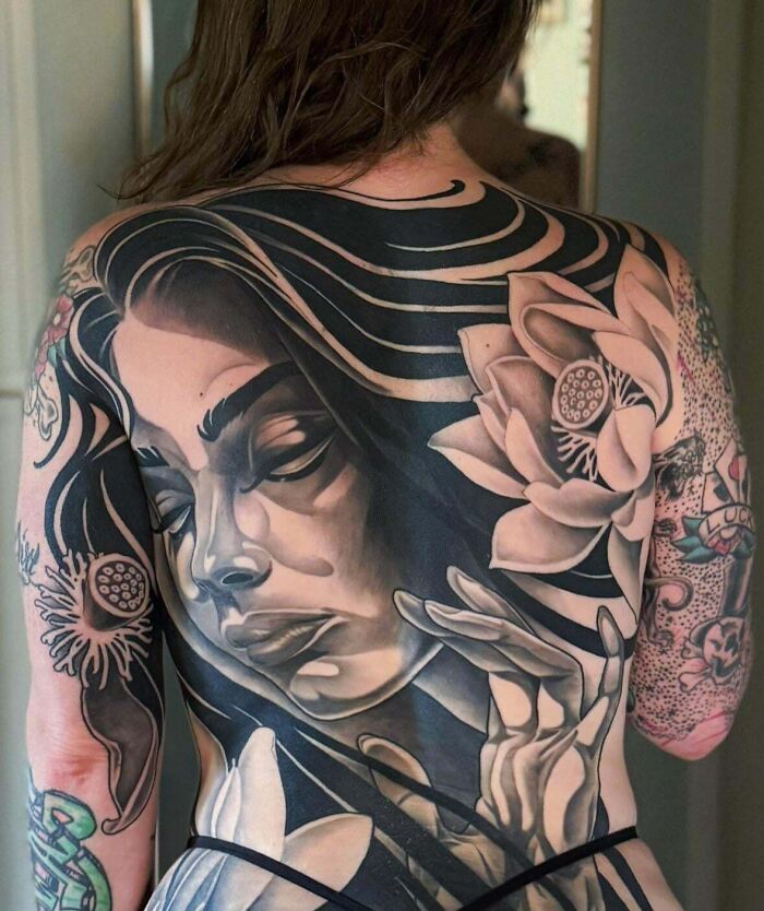 A full back piece tattoos featuring a grayscale portrait of a woman with flowing hair and lotus flowers. Stunning tattoo.