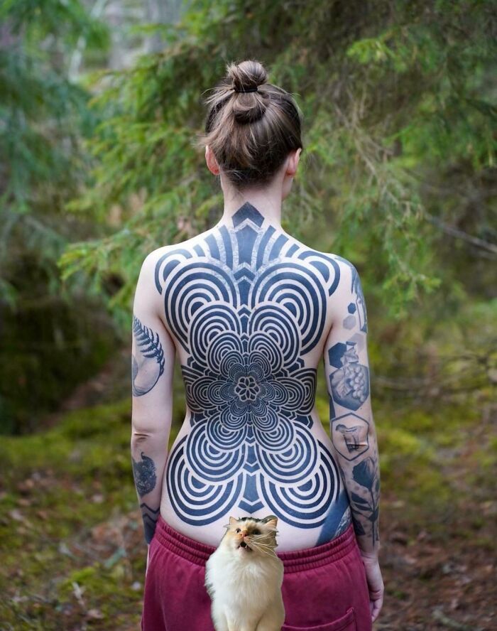 A person with an intricate geometric full back tattoo, standing in a forest, showcasing stunning tattoos.