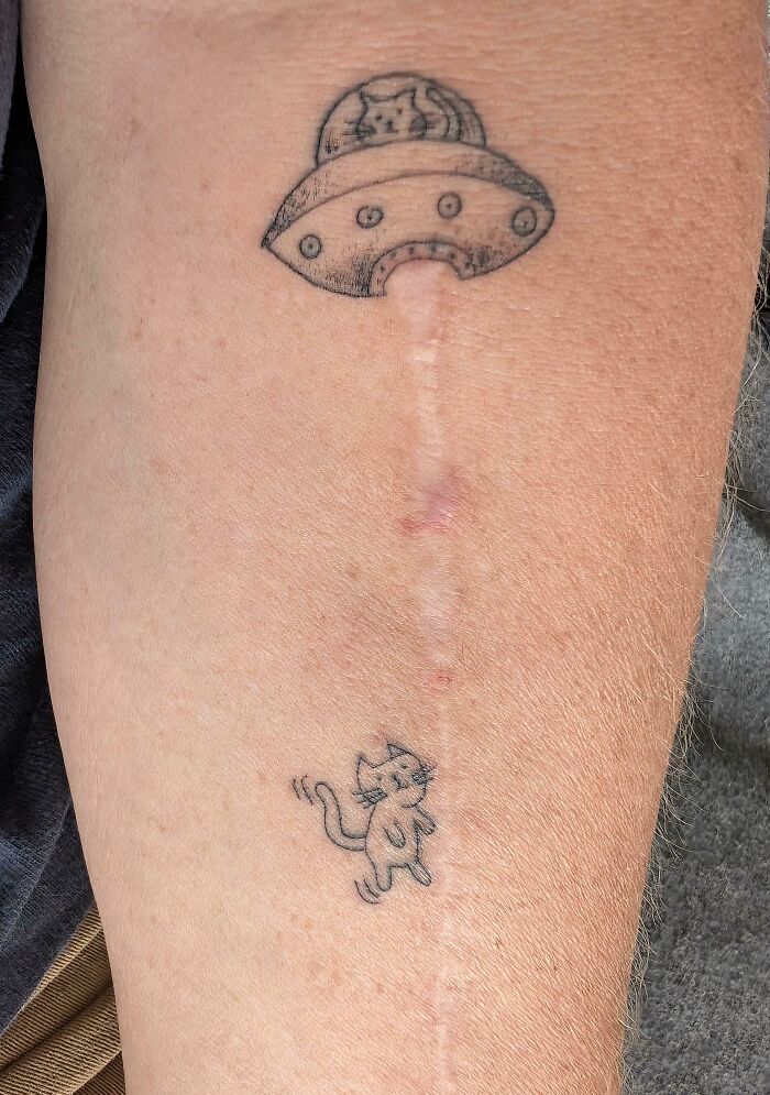 A small, stunning tattoo of a cat being abducted by a UFO, creatively incorporating a scar into the design.