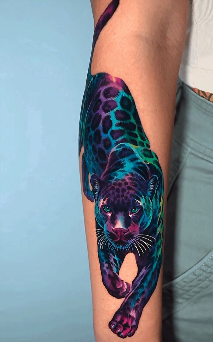A vibrant, colorful leopard tattoo on a person's forearm, featuring shades of purple, blue, and green. This stunning tattoo is permanent.
