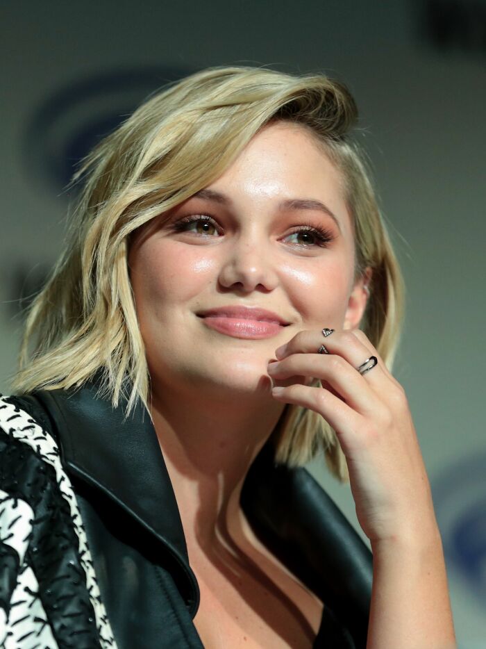 Olivia Holt smiling with short blonde hair and a black and white patterned jacket. Bio and career highlights for Olivia Holt.