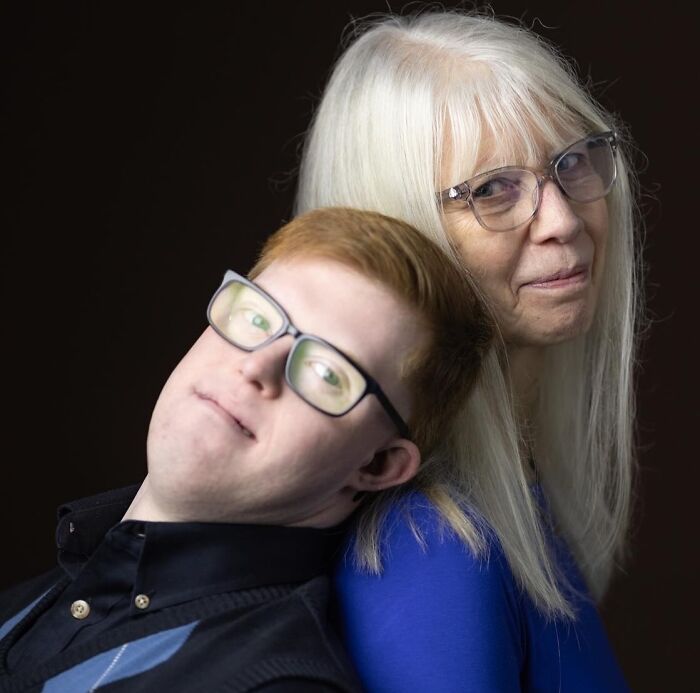 A lovely portrait of a mother with long white hair leaning back-to-back with her natural redhead son, both wearing glasses.