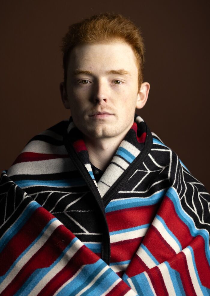 A serious natural redhead man with freckles and a colorful blanket draped over him, from a global portrait project.