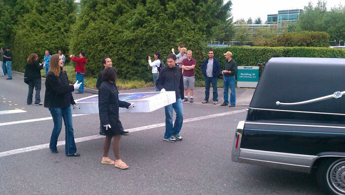 People carrying a large box out of a building, next to a hearse, which aged poorly.
