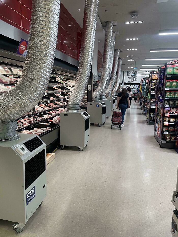 Portable air conditioning units with large silver ducts cooling a grocery store aisle, preventing summer melting.