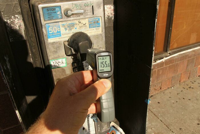 A hand holding a temperature gun showing 155.6 degrees Fahrenheit on a payphone, illustrating summer melting.