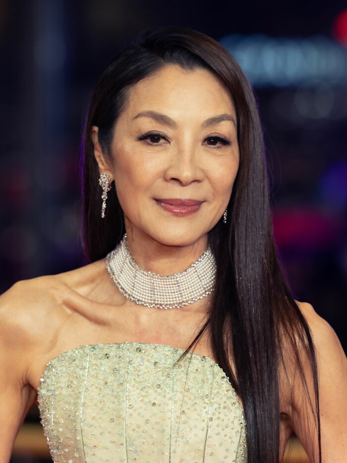 Michelle Yeoh wearing a strapless gown and pearl choker, showcasing her bio and career highlights.