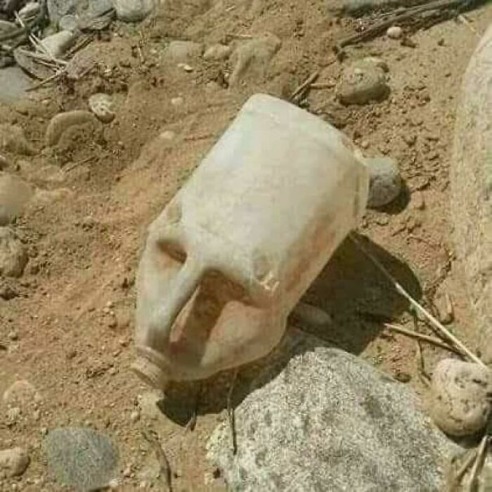 A discarded white plastic container on sandy ground, with a handle that appears to form a human-like nose and face, a confusing pic.