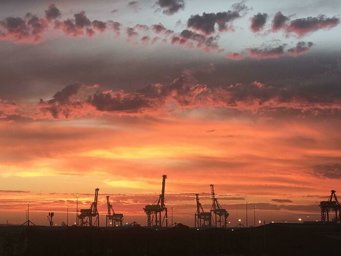 A dramatic sunset over a port, with silhouettes of cranes against a fiery red and orange sky, a confusing pic.