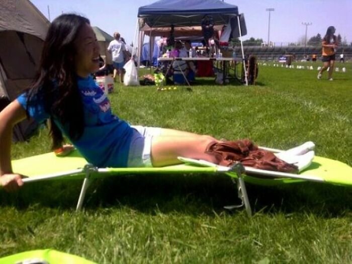 A young woman in a blue shirt resting on a green cot on a grassy field, looking like a confusing pic due to the angle.