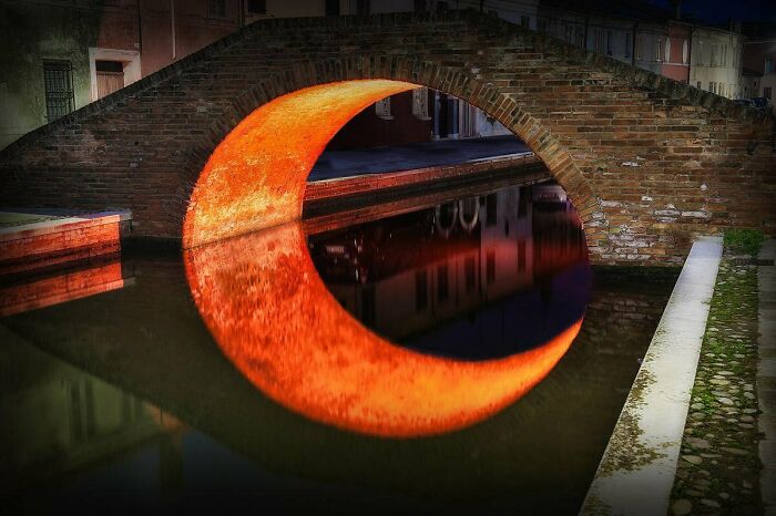 A brick arch bridge over water, reflecting an orange crescent shape, a confusing pic that may melt your brain.