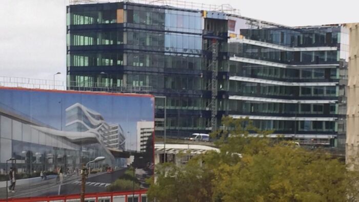 A large billboard displaying an architectural vision of a sleek, wavy building next to the under-construction reality, which looks more angular.