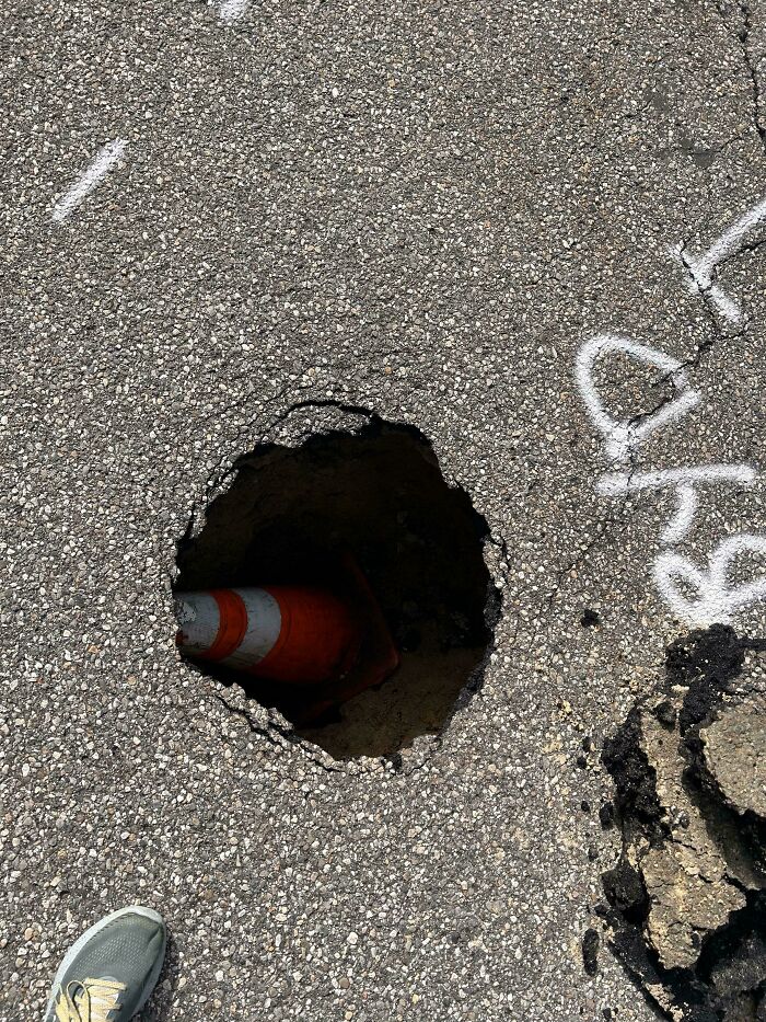 An orange traffic cone has fallen into a large pothole in the asphalt, showing dangerous things people did.