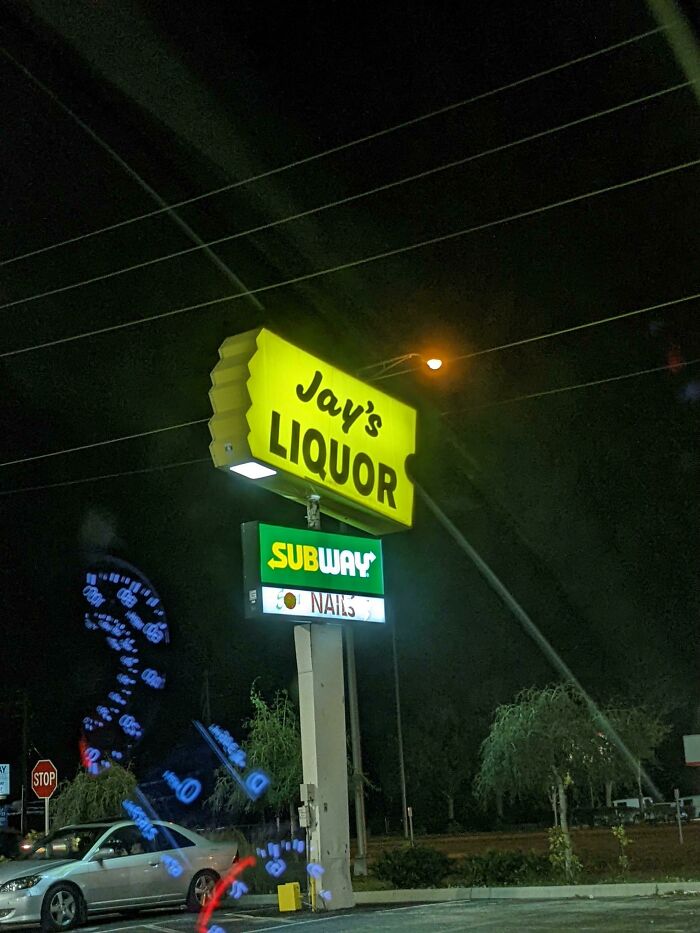 A roadside sign at night for Jay's Liquor, Subway, and Nails, a building pretending to be something different.