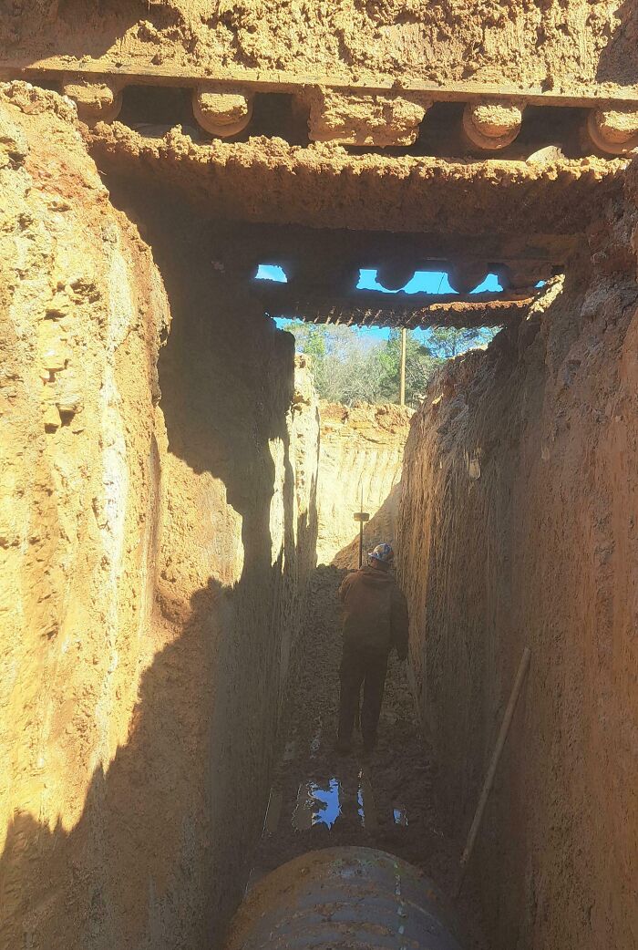 A construction worker in a deep, narrow trench, potentially doing dangerous things, with a large pipe at the bottom.