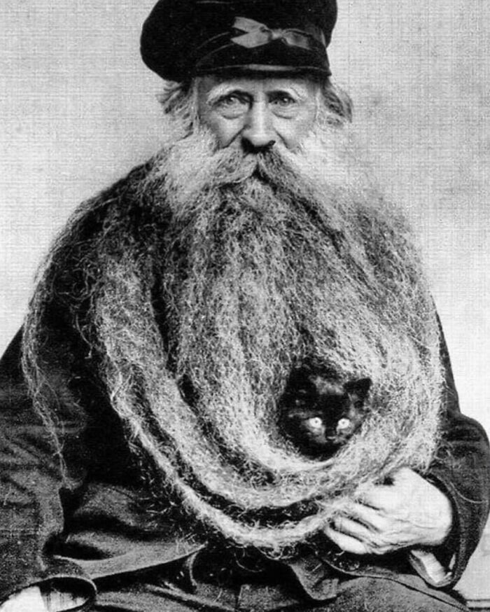 Vintage photo of a man with an extremely long beard, with a small black cat nestled inside. Animals steal the spotlight.