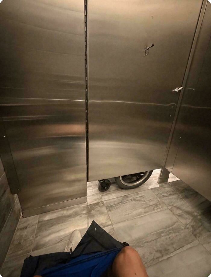 A person using a public restroom with a mobility scooter visible in the stall next to them, one of those pics that people cannot explain.