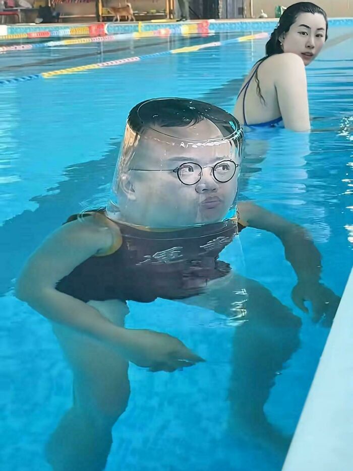 A man in a swimming pool with a clear plastic container over his head, making his face appear distorted, while a woman looks on. A pic people cannot explain.