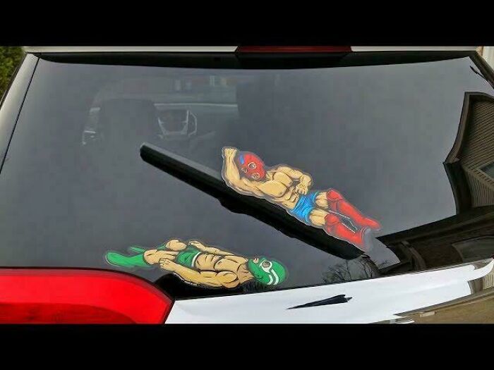Two cartoon wrestler decals are placed on a car's rear window, positioned to interact with the wiper, a pic people cannot explain.