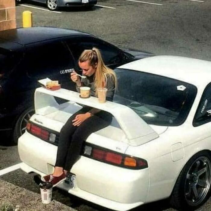 A woman sits on the spoiler of a white car, using it as a table to eat a meal, a pic people cannot explain.