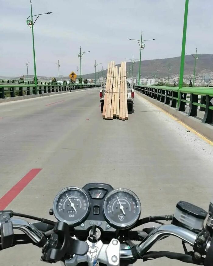 A motorcycle rider's view of a truck on a bridge, carrying an inexplicably tall and unsecured load of wood.