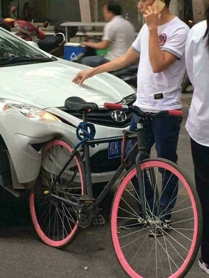A bicyclist on a phone stands next to a white car that has its front fender smashed by a bicycle, an unexplainable pic.