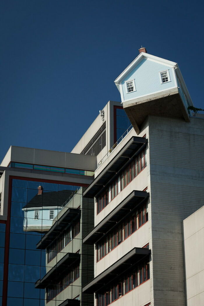 A small house precariously perched atop a modern building with a reflection in the glass, a pic that people cannot forget.