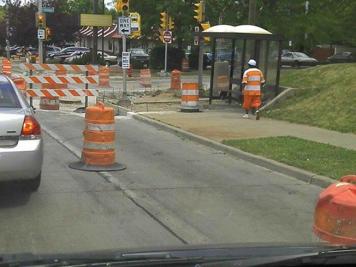 A man in orange uniform walks past road construction barrels and a bus stop, a pic people cannot explain.