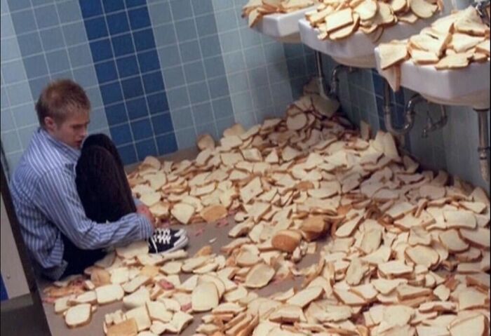 A person sitting in a bathroom with the floor covered in bread slices, an unexplainable pic that people cannot explain.