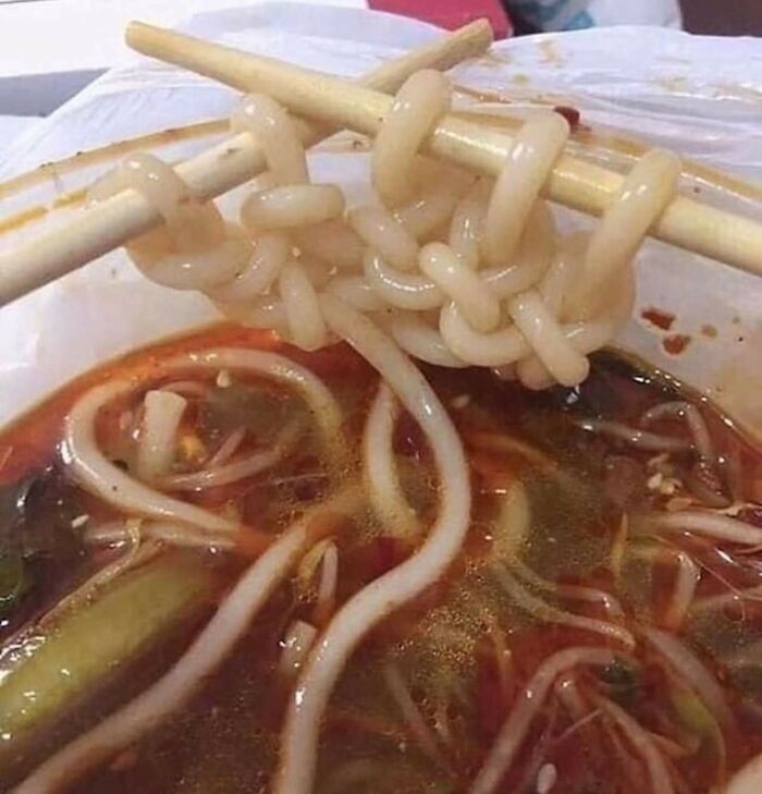Chopsticks hold unusually thick, tied-together noodles over a bowl of soup, an unexplainable food moment.
