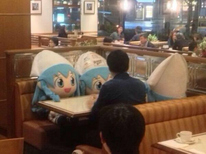 Two large mascot heads of anime characters sit in a restaurant booth with a person, an unexplainable sight.