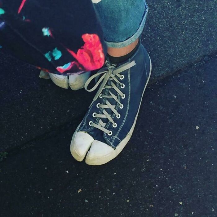 A person wears denim jeans and unique split-toe canvas shoes, one foot visible, on dark pavement.