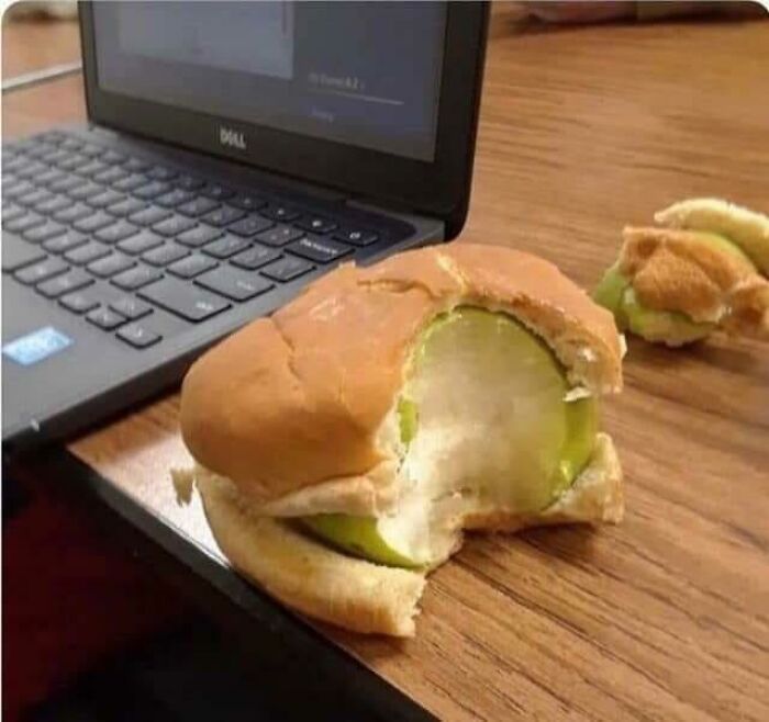A bitten green apple inside a burger bun next to a laptop on a wooden desk. Cannot Explain Pics.