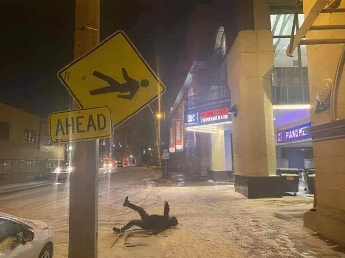 A person fallen on a snowy sidewalk next to a tilted pedestrian crossing sign, one of those pics that people cannot explain.