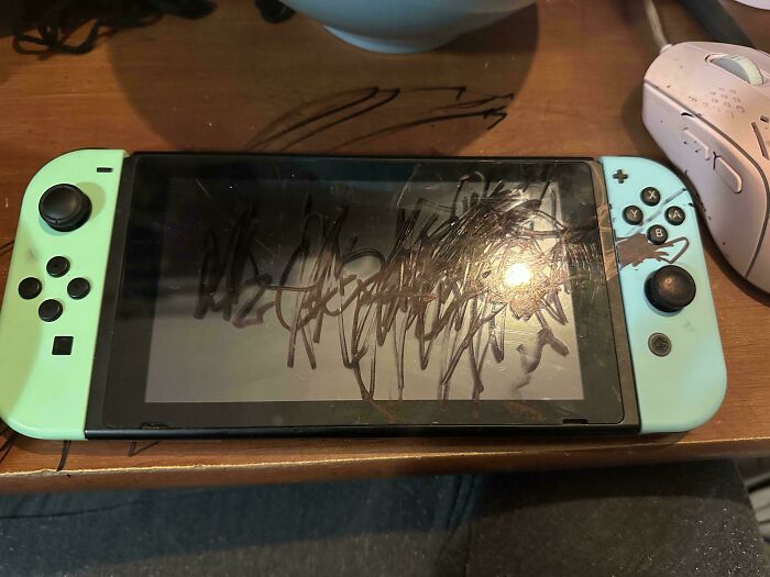 A close-up of a light green Nintendo Switch console with its screen and joy-cons heavily scribbled on with a dark marker, showing parents' struggles.