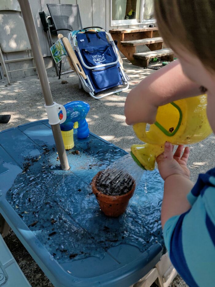 A young child watering a small plant in a pot on a blue table, creating a mess, making parents wonder why they signed up for this.