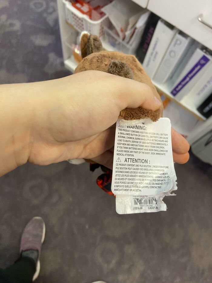 A hand holding a small toy with a warning label about button cell batteries, reminding parents of safety concerns.