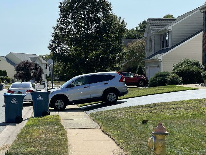 A silver SUV poorly parked on a driveway with its front wheels on the sidewalk, showcasing jerk neighbors parking issues.
