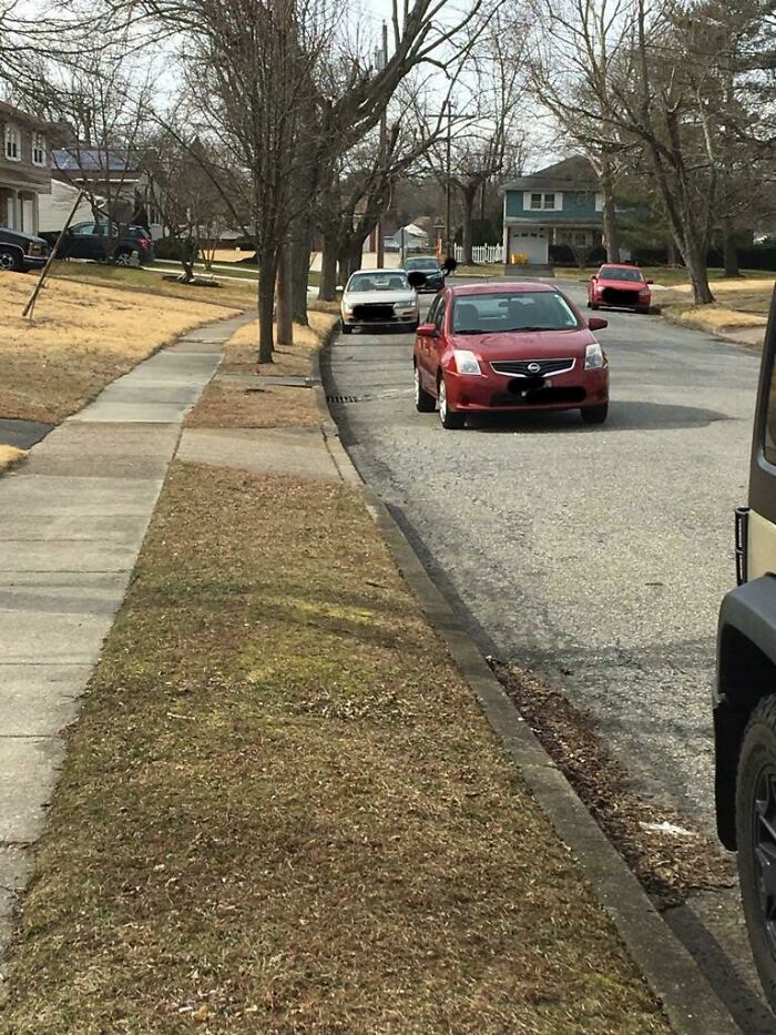 A red car, a prime example of jerk neighbors parking, is parked in the street partially blocking the flow of traffic.