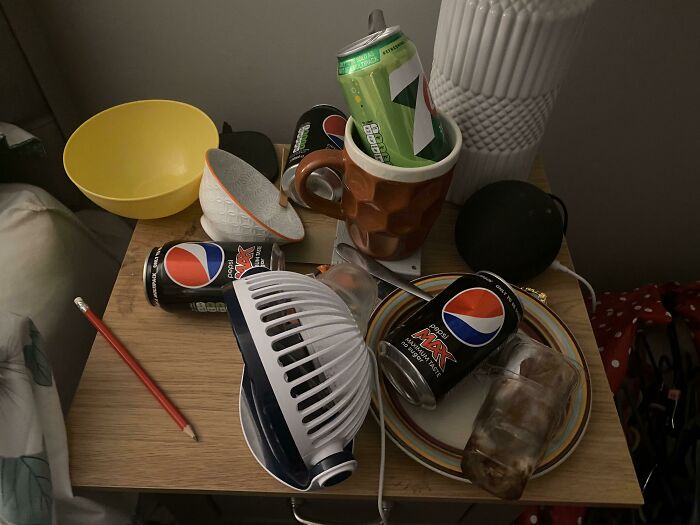 A cluttered bedside table with empty soda cans and various items, making becoming a cat lady look more attractive.