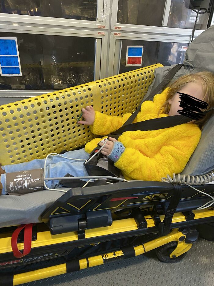A child in a yellow fluffy coat and jeans lies on a stretcher inside an ambulance, with monitoring equipment on their arm.