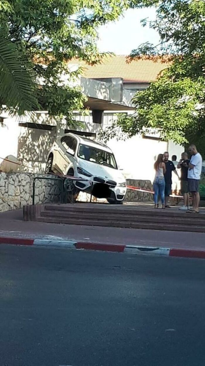 A white SUV in an urgent parking lesson, precariously perched on a stone wall above steps, watched by neighbors.