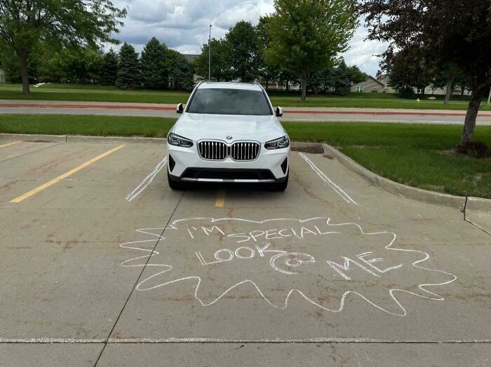 A white car poorly parked in a parking spot with a chalk outline and text, I'm special look @ me.