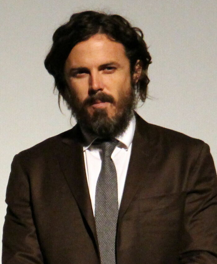 Casey Affleck with dark, shaggy hair and a full beard, wearing a brown suit and grey tie, highlighting his career highlights.